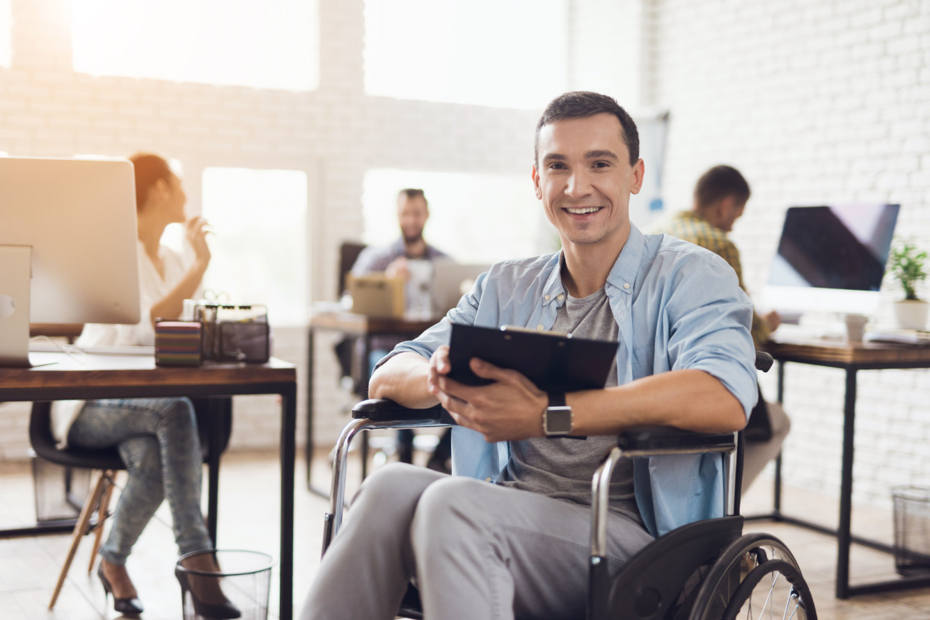 A person in a wheelchair holding a tablet smiles in a bright, modern office with colleagues working in the background.