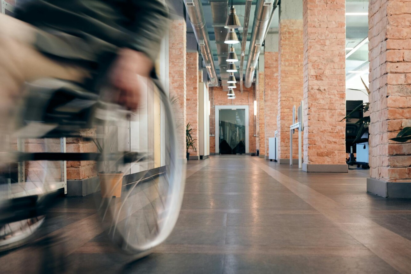 A modern office hallway with exposed brick walls and a blurred person in a wheelchair in the foreground.
