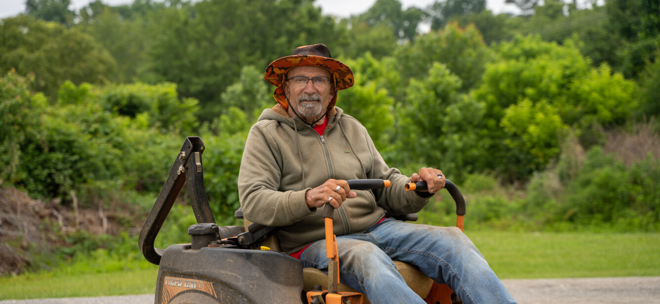 Rick Motes is a white middle-aged man with an orange hat, grey goatee, wearing glasses. He is seated in a lawn mower.