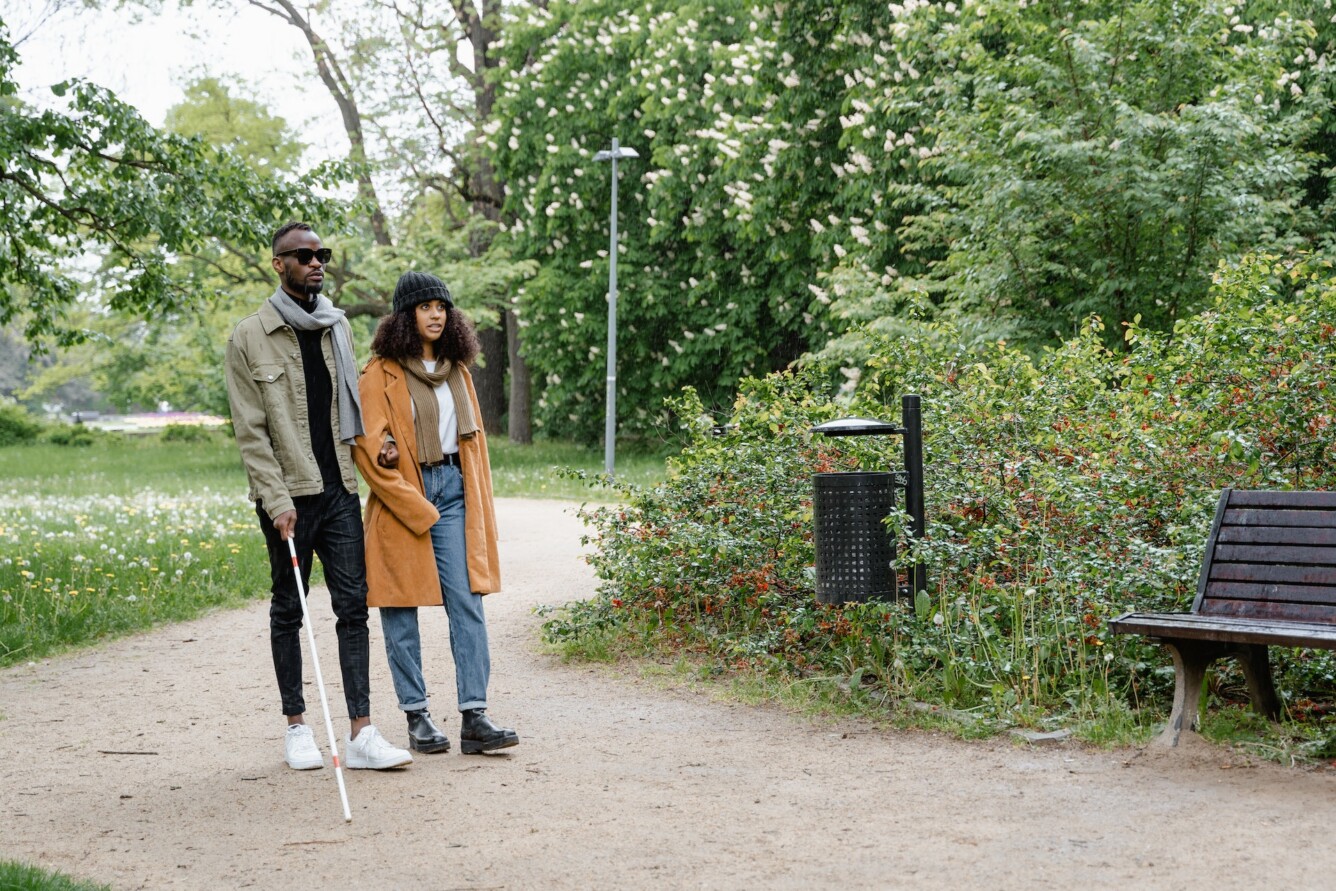 Two individuals walk side by side on a gravel trail lined with grass and bushes. The person on the left wears sunglasses and holds a white cane, walking arm-in-arm with the person on the right.