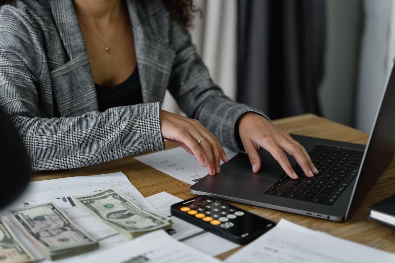 Person using a laptop at a table with documents, a smartphone calculator, and dollar bills.