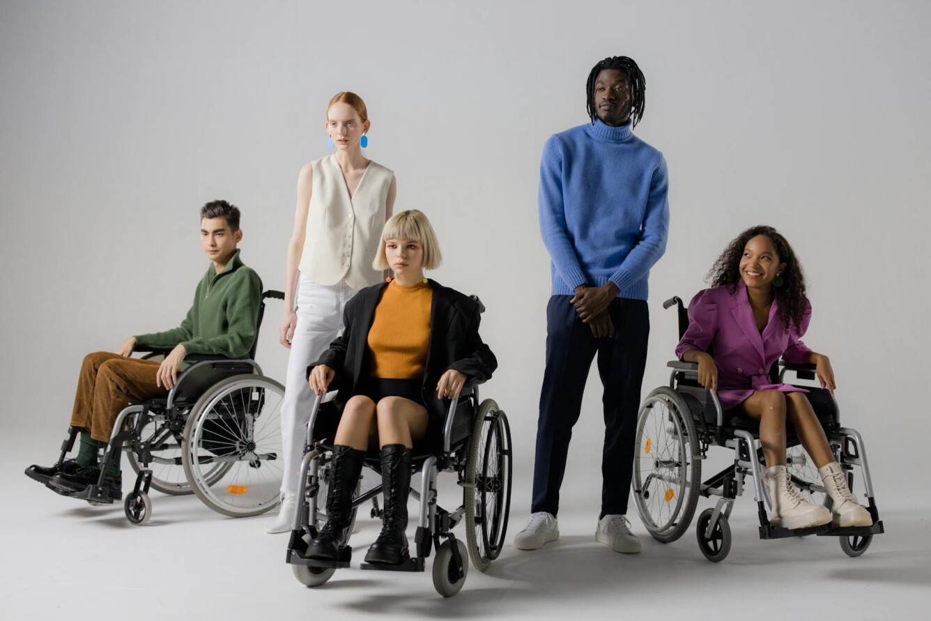 Five individuals, three in wheelchairs, in a neutral setting; diverse attire and postures.