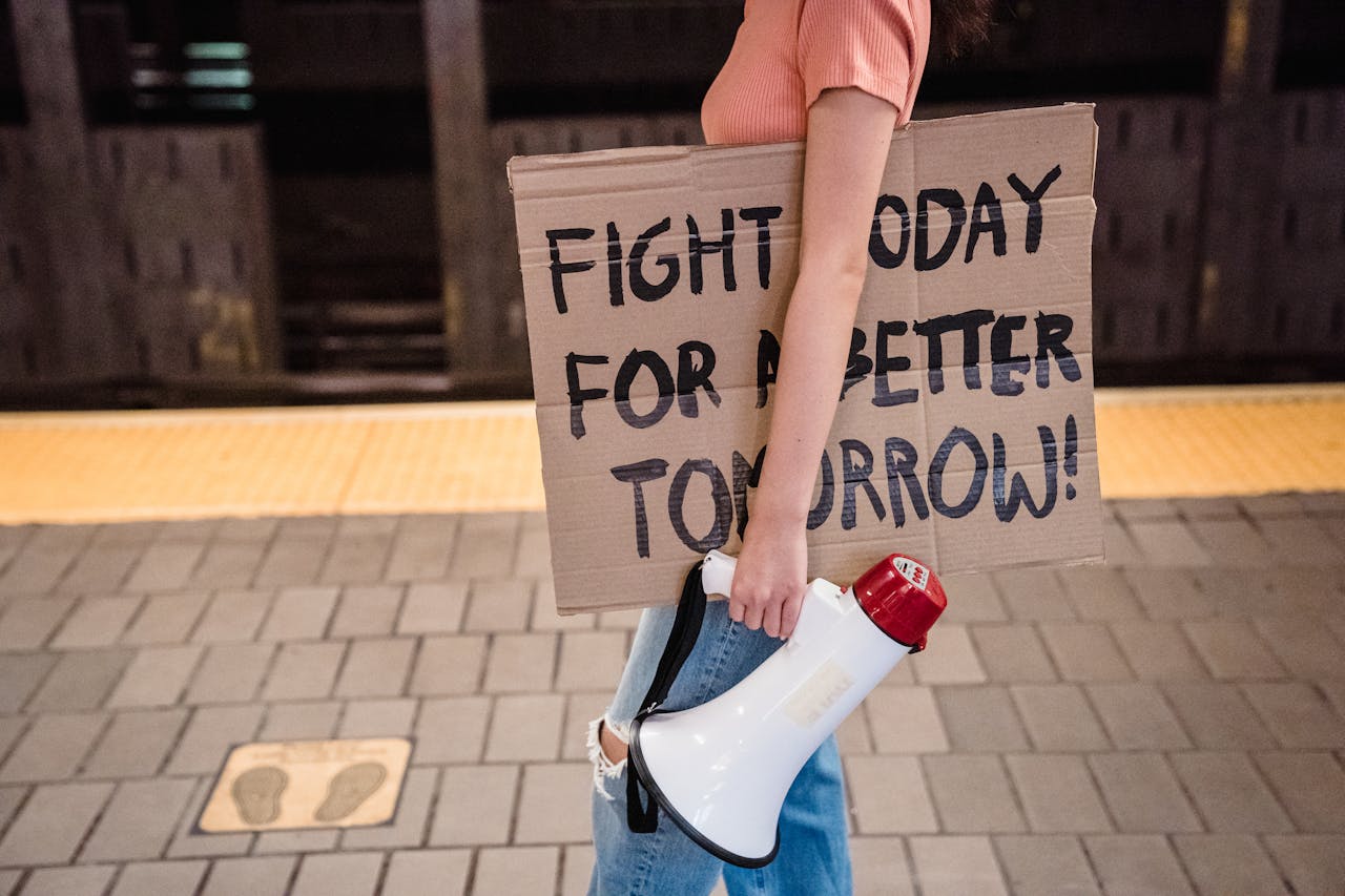 Person holding a megaphone and a cardboard sign on a subway platform. Transcribed Text: "FIGHT TODAY FOR A BETTER TOMORROW!"