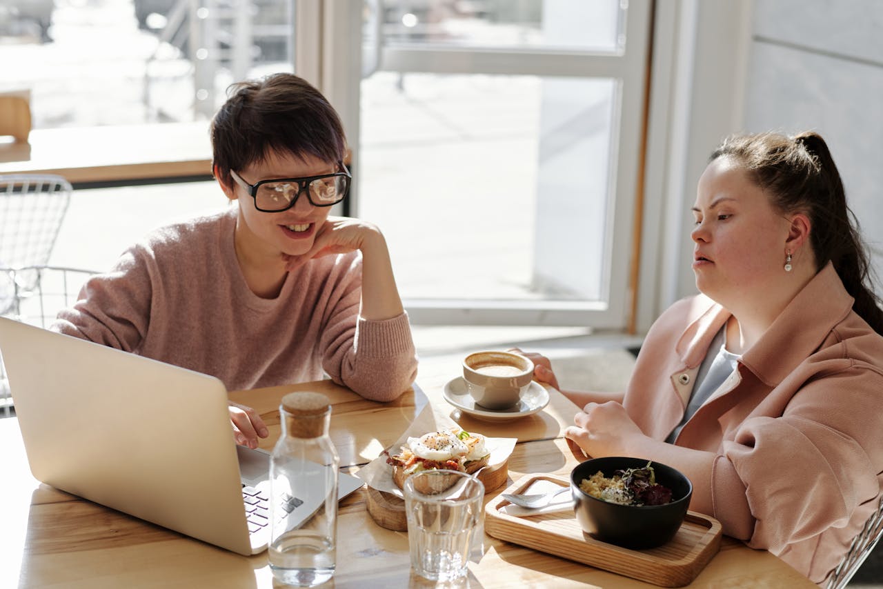 Two people sitting at a table with a laptop, coffee, and food in a sunlit room.