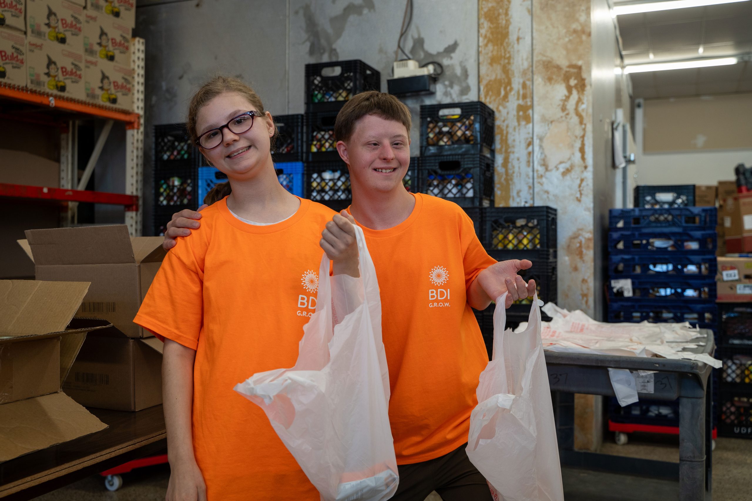 A girl and a boy wearing orange GROW shirts smiling and holding white plastic bags volunteering at a local food bank.