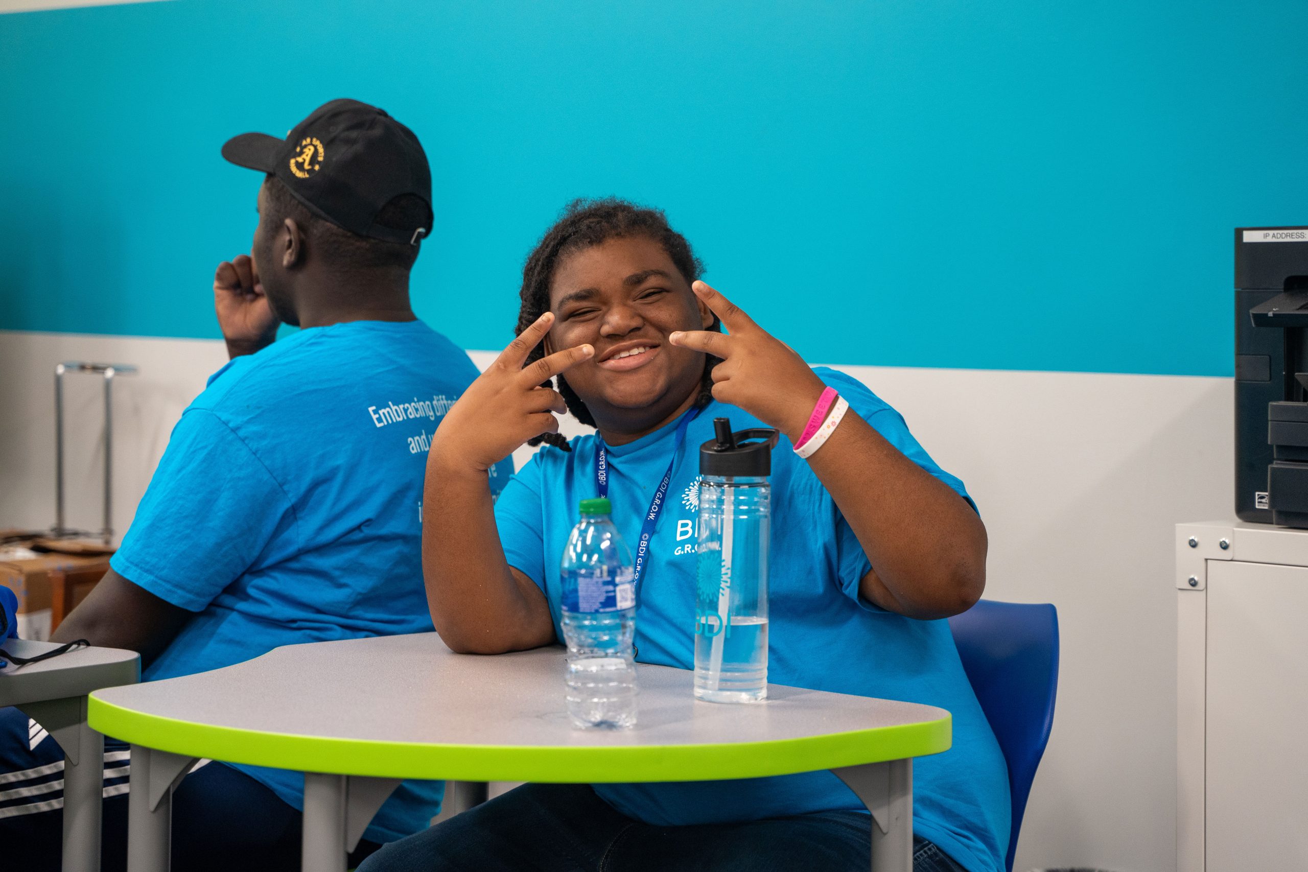 A GROW participant smiling and making peace signs at a table with water bottles in a classroom.