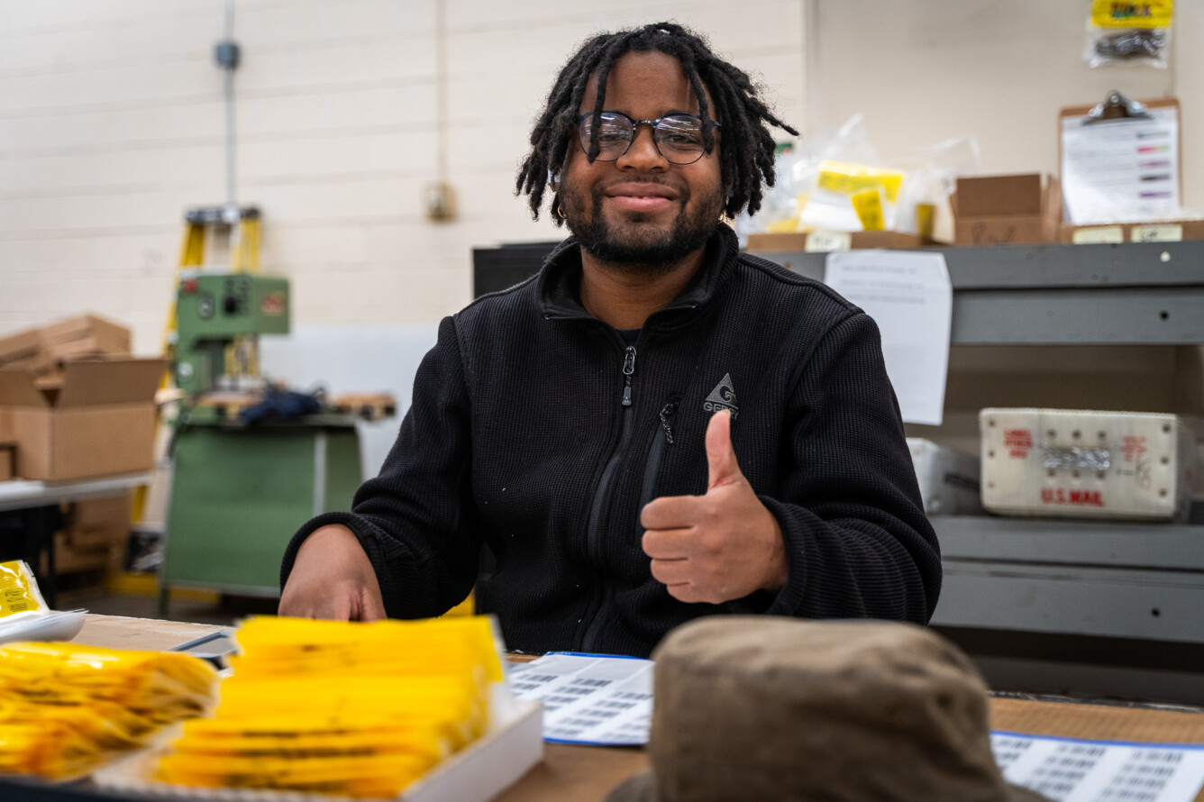 Todd Willis, BDI production worker, at his work desk in the Atlanta warehouse. He is smiling and showing a thumb up.
