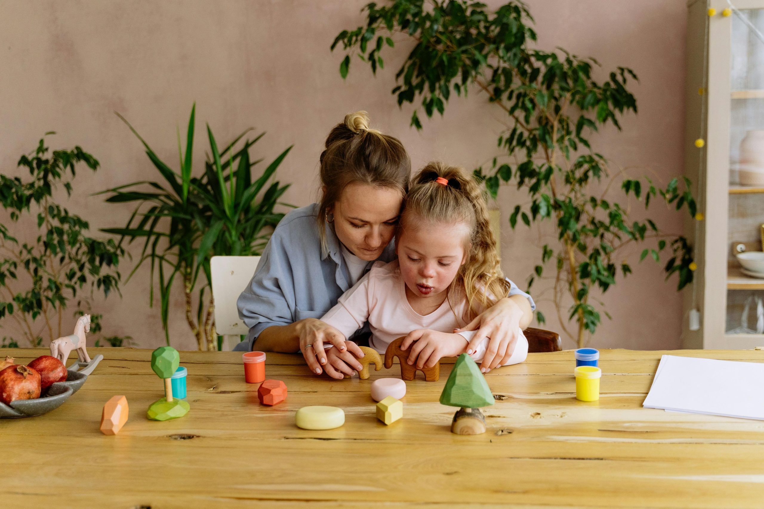 Woman and girl playing with toys at a wooden table, surrounded by colorful blocks and plants in the background.