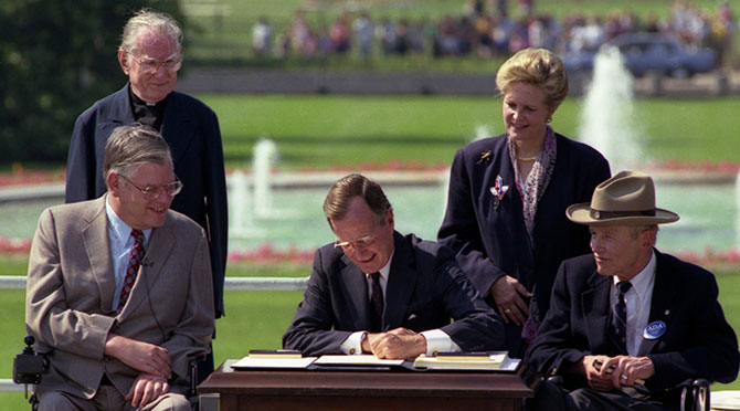 President George Bush signs into law the Americans with Disabilities Act of 1990 on the South Lawn of the White House. L to R, sitting: Evan Kemp, Chairman, Equal Employment Opportunity Commission, Justin Dart, Chairman, President's Committee on Employment of People with Disabilities. L to R, standing: Rev. Harold Wilke and Swift Parrino, Chairperson, National Council on Disability