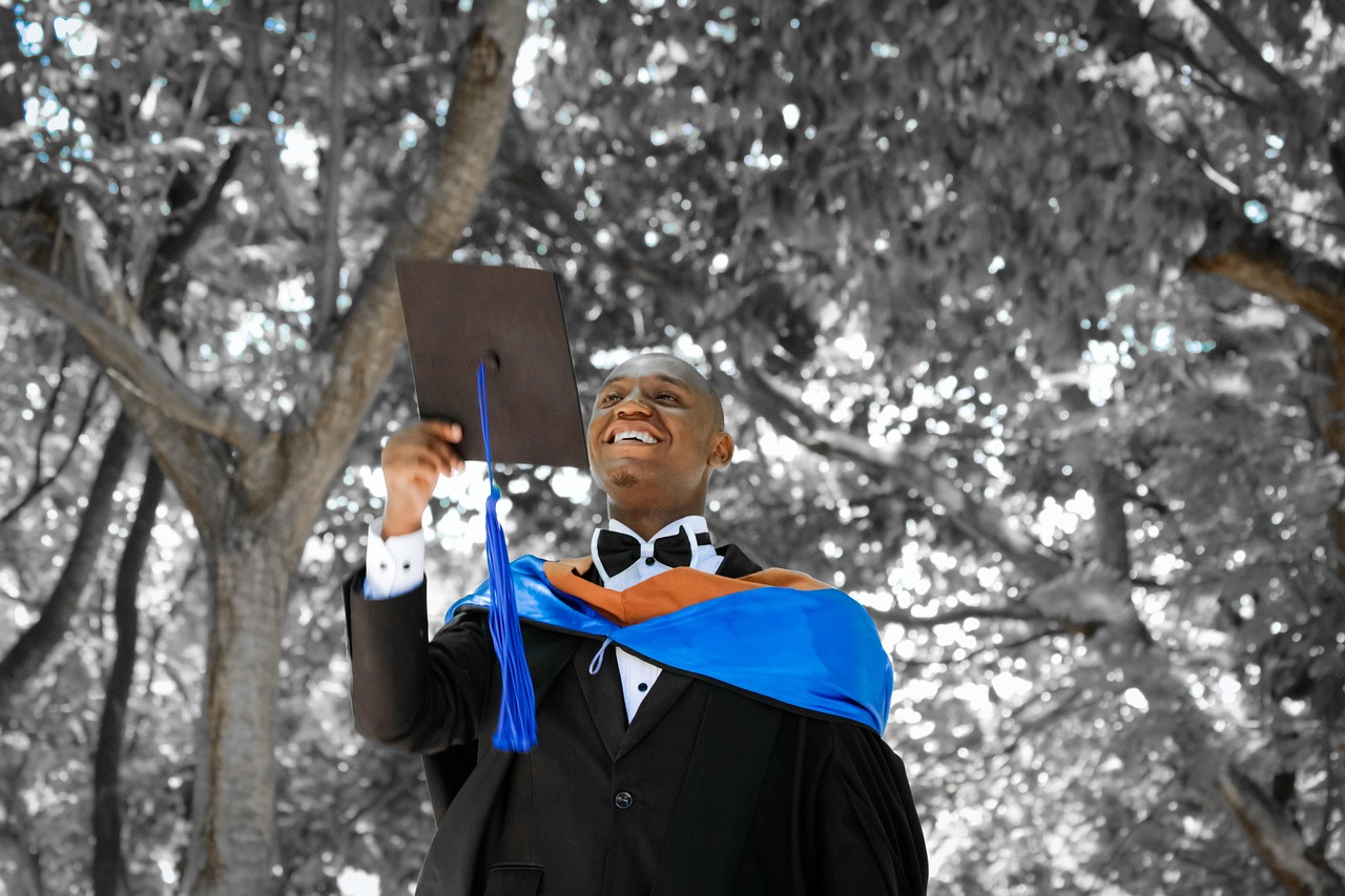 A graduate holding up a graduation hat, wearing a black gown with a blue and orange stole, smiling under a tree canopy.
